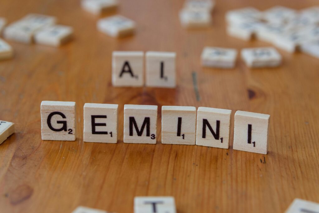 Scrabble tiles spell out the words 'Gemini' and 'AI' on a wooden surface,